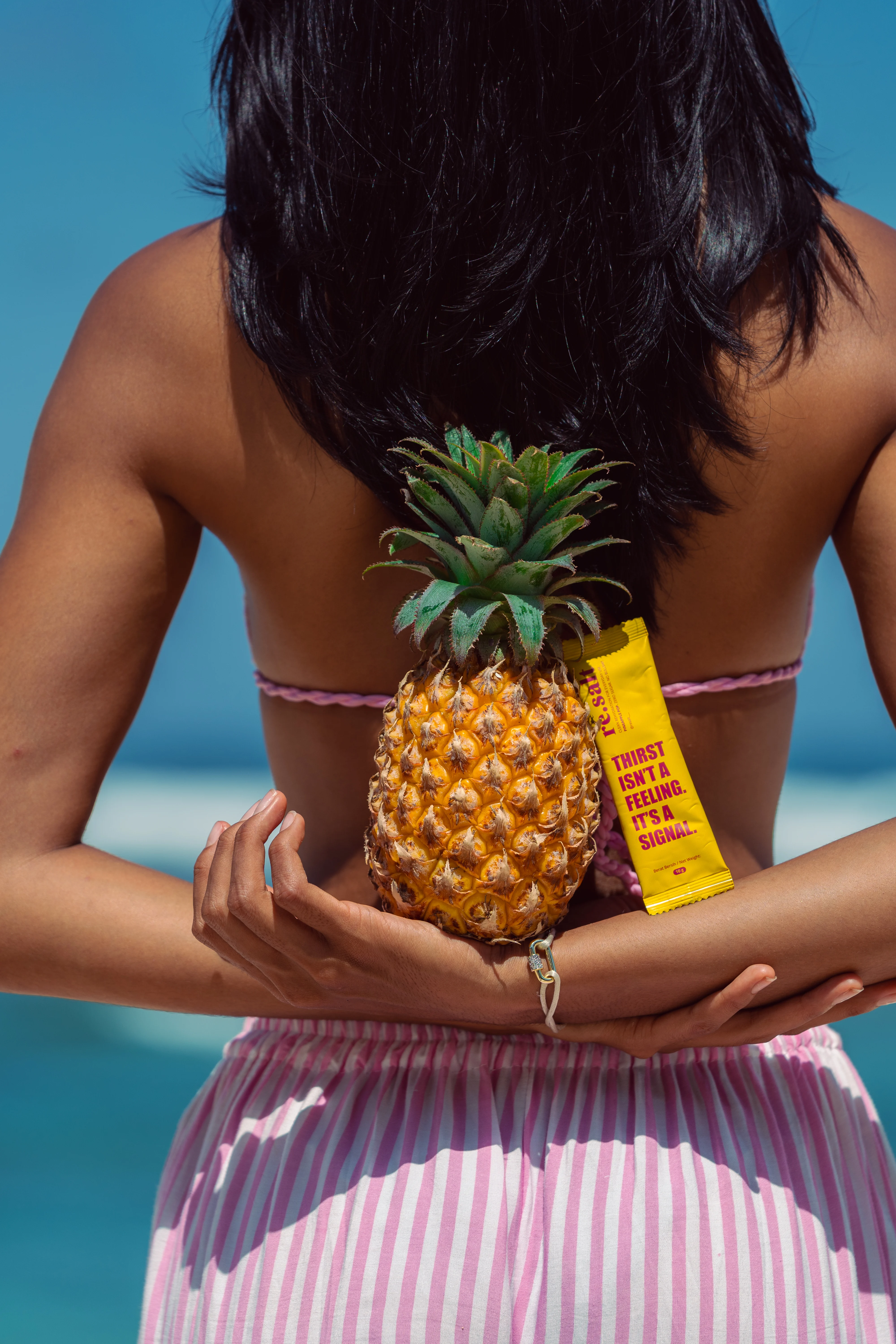 Woman seen from behind at the water's edge, holding a whole pineapple and a yellow re.salt sachet.