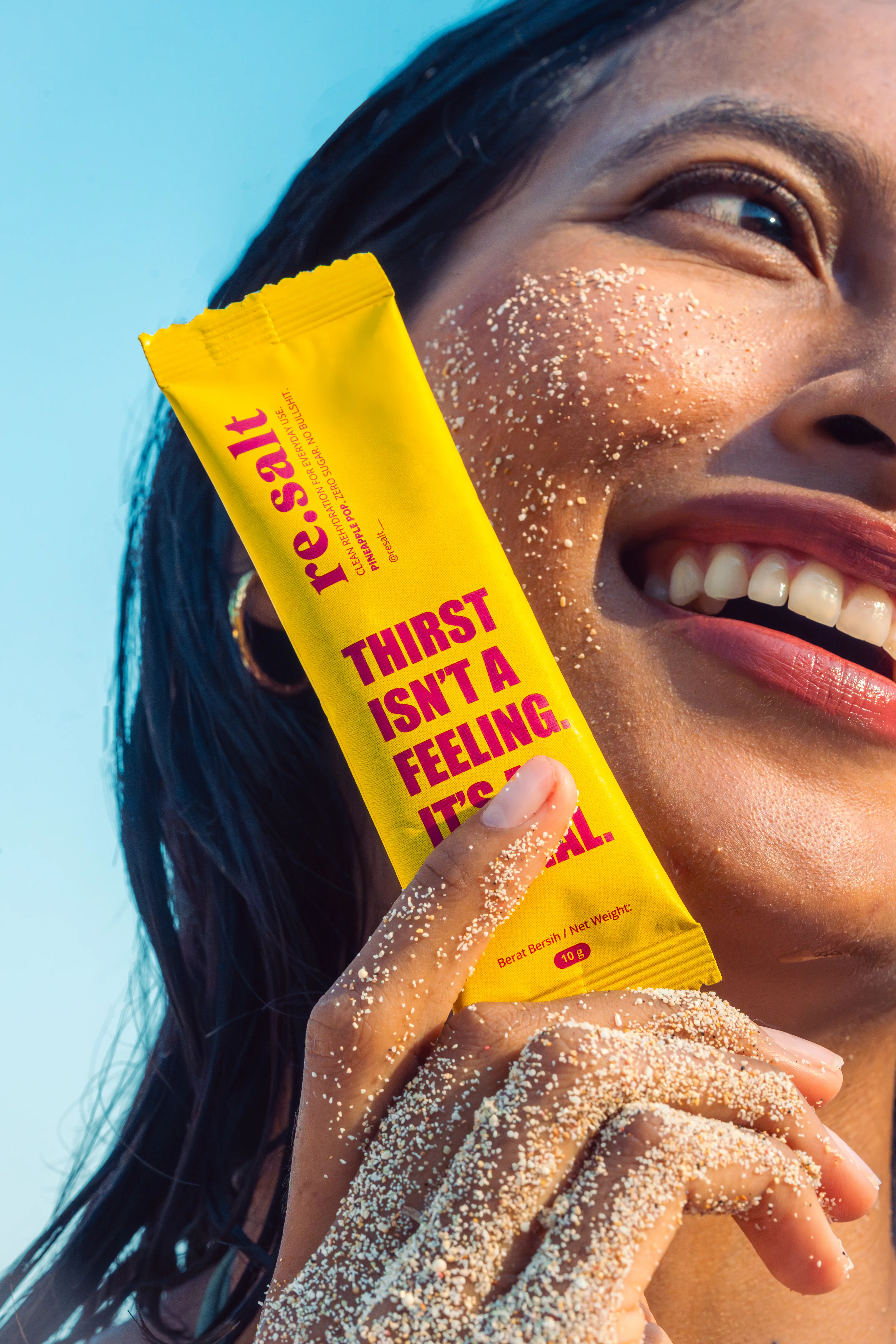 Smiling woman holding a Pineapple Pop sachet against her sandy cheek